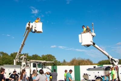 People standing on the ground while two buckets from line trucks are up in the air opposite each other with a lineman and child in each bucket