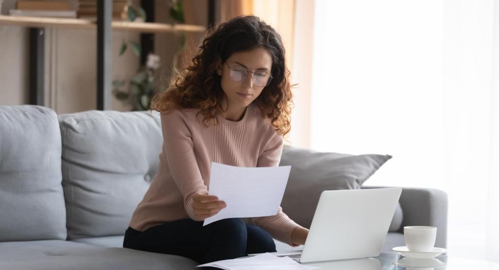 Woman sitting on couch in front of laptop computer holding paper